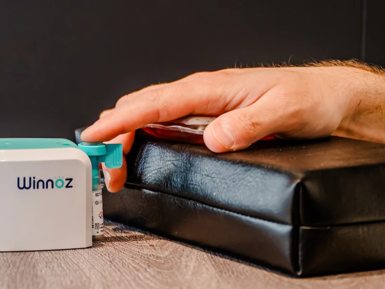 A hand rests on a black cushion next to a medical device labeled "Winnoz," with a finger touching a teal button on the device.