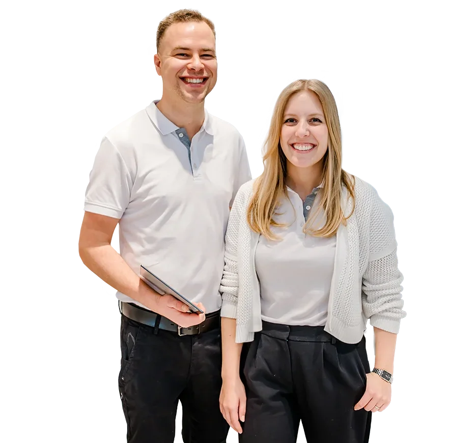Two people are smiling and standing side by side. They are wearing casual attire with light-colored shirts. One person is holding a tablet. The background is plain.