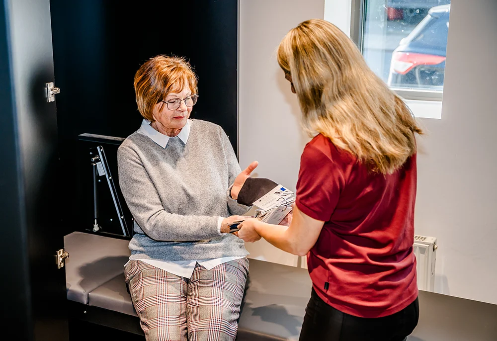A woman in a gray sweater and plaid pants sits on a bench while another woman in a red shirt hands her a device. They are indoors near a window.