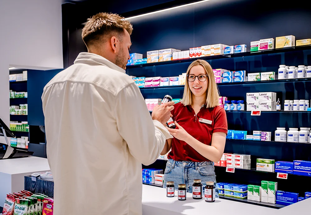 A pharmacist in a red shirt assists a customer at a pharmacy counter. Shelves behind them are stocked with various medicines and supplements. The customer, wearing a white jacket, holds a bottle.
