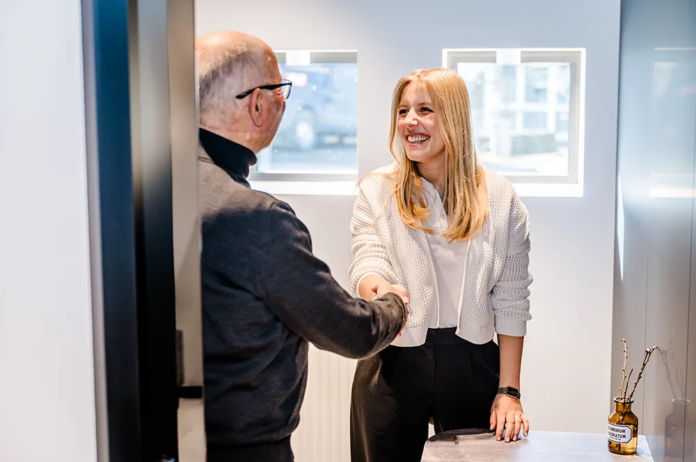A woman with blonde hair wearing a white sweater is smiling and shaking hands with a bald man in a black sweater at an office door. A small vase with dried branches sits on a table nearby.