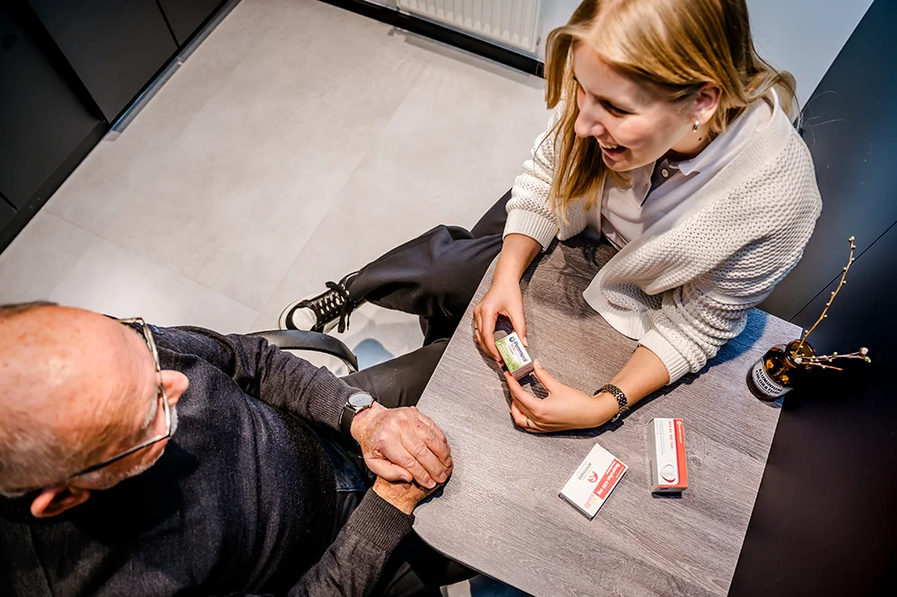 A woman and an older man are sitting at a table, holding hands. The woman is smiling and holding a medicine box. Several other medicine boxes are on the table. They are in a room with a gray floor and dark furniture.