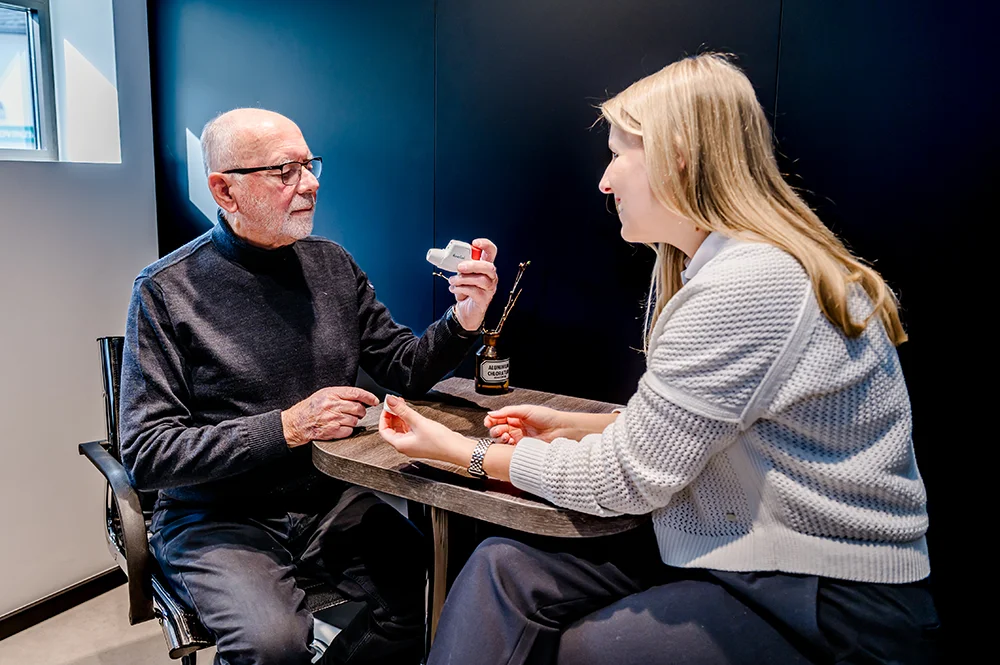 A man and a woman sitting at a table in a modern room. The man, wearing glasses and a dark sweater, is holding a dental model. The woman, dressed in a white sweater, is smiling and interacting with him. A small window is in the background.