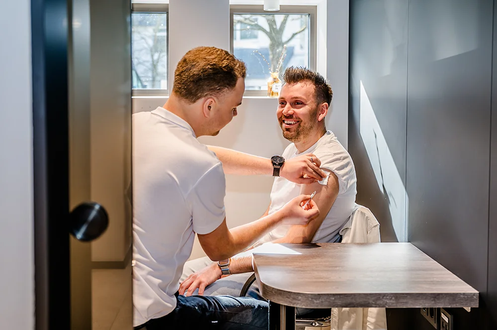 A healthcare professional administers a vaccine to a smiling man sitting in a chair. The setting appears to be a clinical room with a window in the background. Both individuals are casually dressed.