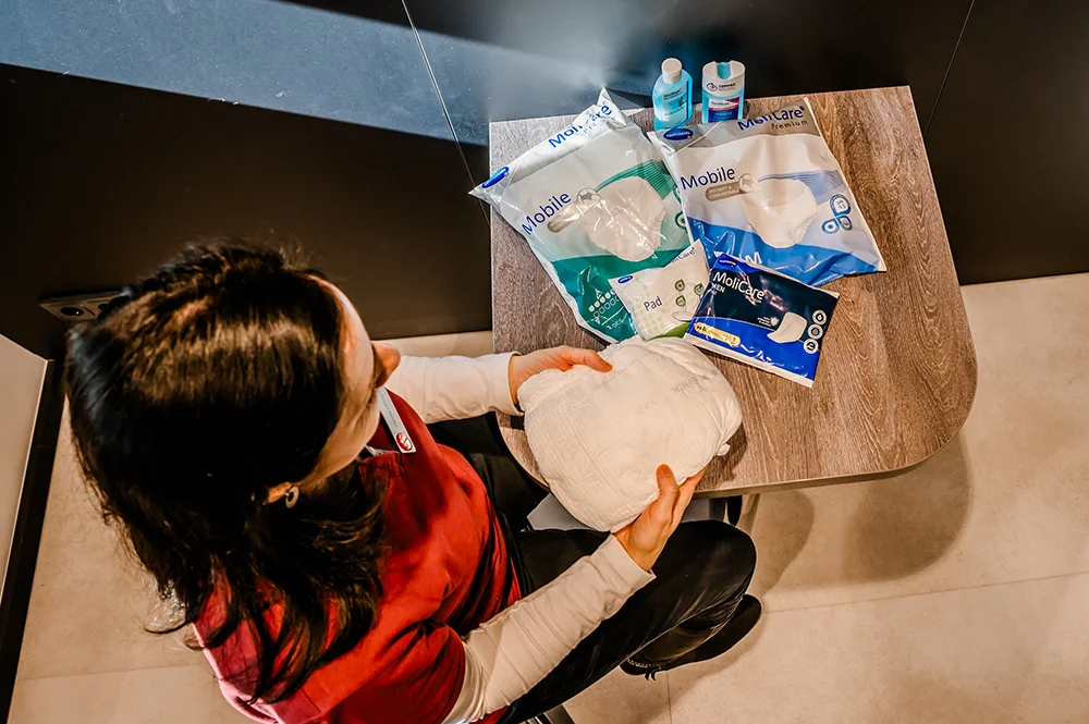 A person in a red shirt is seated at a table, holding an incontinence pad. The table has various incontinence products, including packages labeled "Mobile," alongside two bottles of hygiene products.