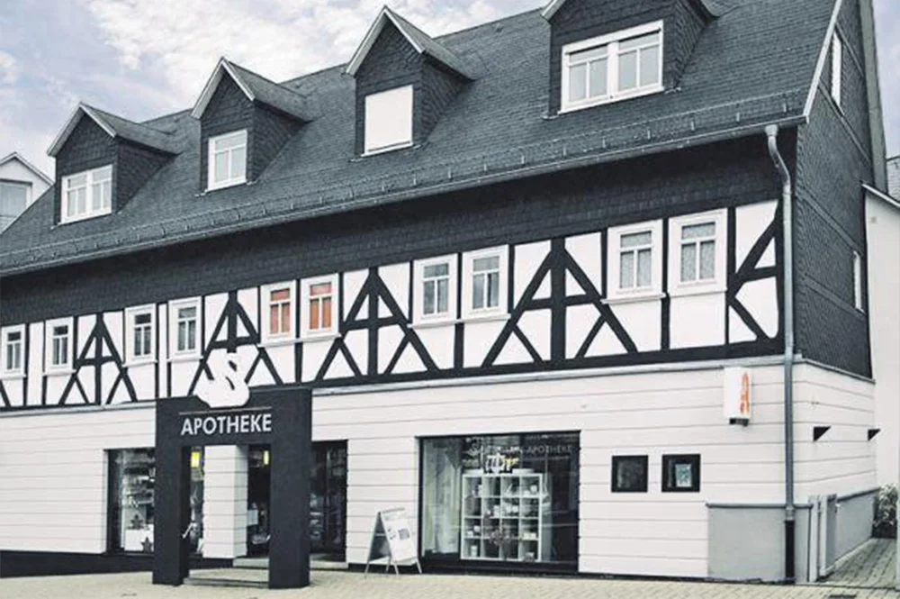 A traditional half-timbered building with a gray roof houses an "Apotheke," or pharmacy. The exterior is white with contrasting dark timber accents. The front features a black sign and a sidewalk sandwich board.