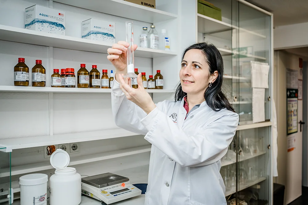 A woman in a lab coat examines a test tube in a laboratory. Shelves with bottles and lab equipment are visible in the background. She appears focused on her work.