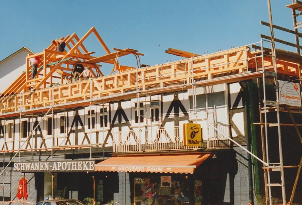 Workers are constructing a timber-framed roof on a building with storefronts below. Signs read "SCHWANEN-APOTHEKE" and "BÄCKEREI SAHN" with pastries displayed. Scaffolding is visible, and the sky is clear.