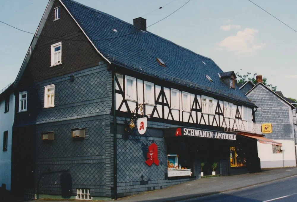 A traditional German half-timbered building with a slate roof houses the Schwanen-Apotheke pharmacy. The exterior features white-framed windows and a red pharmacy symbol. Located on a street corner, another building is visible nearby.