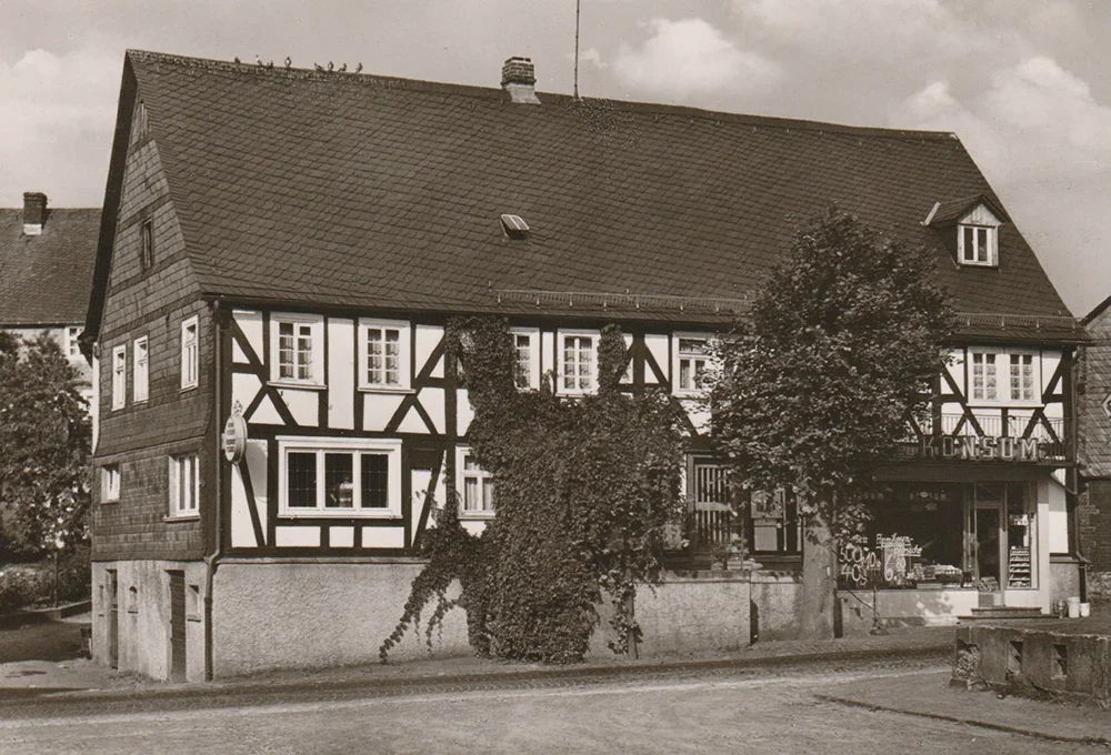 A vintage photograph of a traditional half-timbered house with ivy growing on the facade. The building has a steeply pitched roof and several small windows. A tree stands next to it, and there is cobblestone pavement in front.