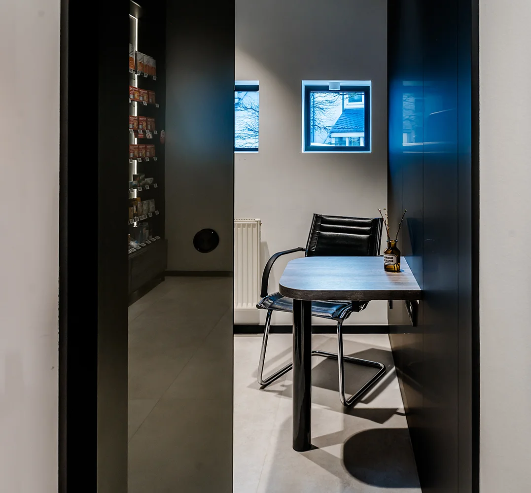 Modern, minimalist room with a small table and two sleek chairs near a window. Shelves with skincare products are visible on a wall in the background. Natural light streams through the window, casting soft shadows on the floor.