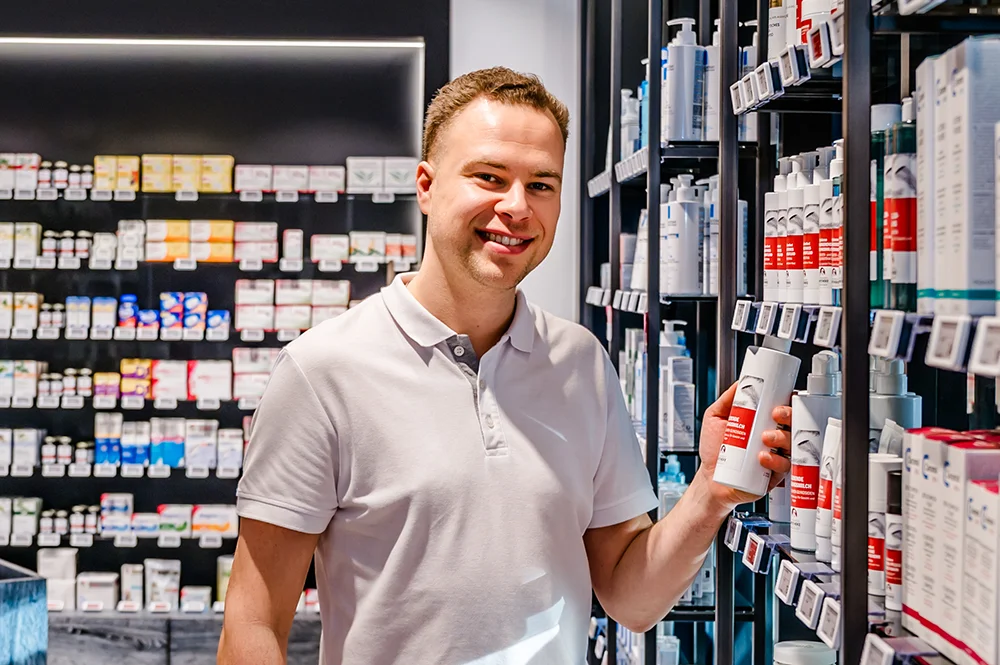 A person wearing a white polo shirt smiles while holding a skincare product in a store aisle. Shelves filled with various boxed and bottled products are visible in the background.
