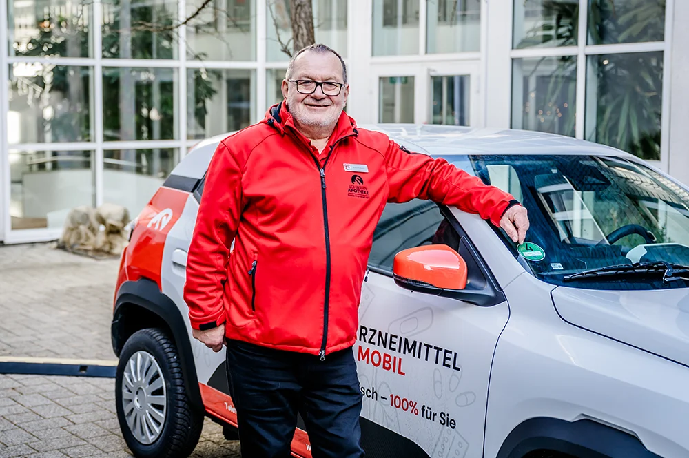 A person in a red jacket stands smiling next to a car with promotional text. The car appears to be parked outside a building with large windows and plants visible inside.