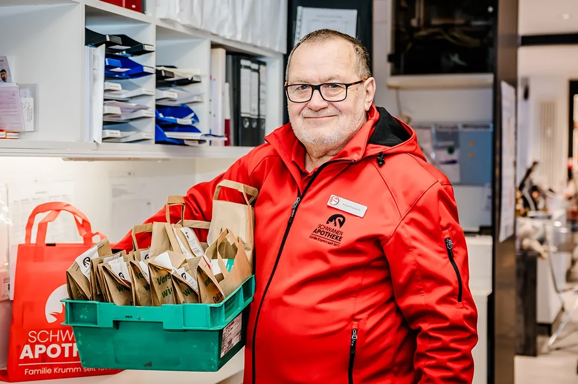 A man wearing a red jacket stands in a room with shelves filled with files. He is holding a green crate containing paper bags. The jacket and nearby items have pharmacy logos on them.