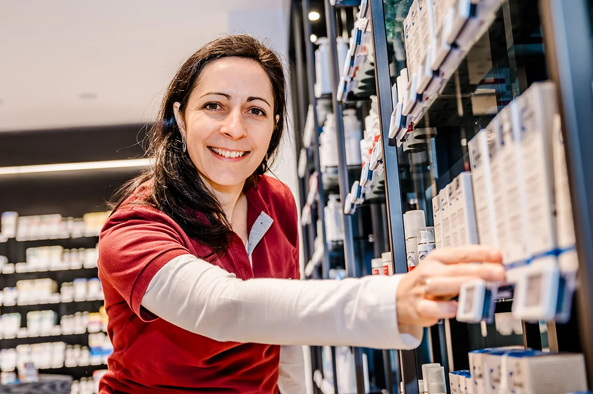 A person wearing a red and white shirt is selecting items from a shelf in a store. They are smiling and appear to be in a pharmacy or retail environment. The background shows additional shelves filled with various products.
