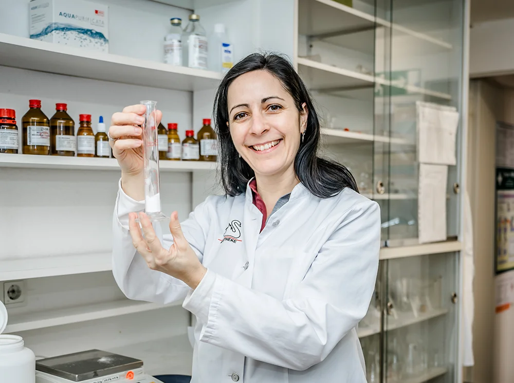 A woman in a lab coat smiles while holding a test tube in a laboratory. Shelves with bottles and scientific equipment are in the background. She appears engaged and focused on her work.