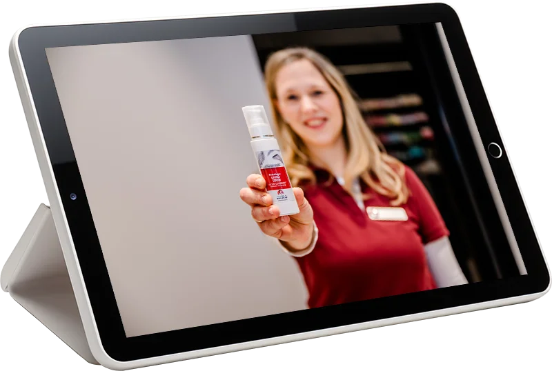 Auf dem Bildschirm eines Tablets ist eine Frau in einem roten Hemd zu sehen, die lächelt und eine kleine Sprühflasche mit rotem Etikett in die Kamera hält, als würde sie sie dem Betrachter anbieten.
