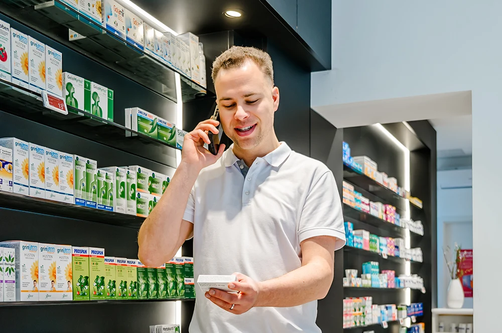 Ein Mann im weißen Poloshirt steht lächelnd in einer Apotheke, telefoniert, hält eine Medikamentenpackung in der Hand und blickt auf Regale voller Medikamentenpackungen.