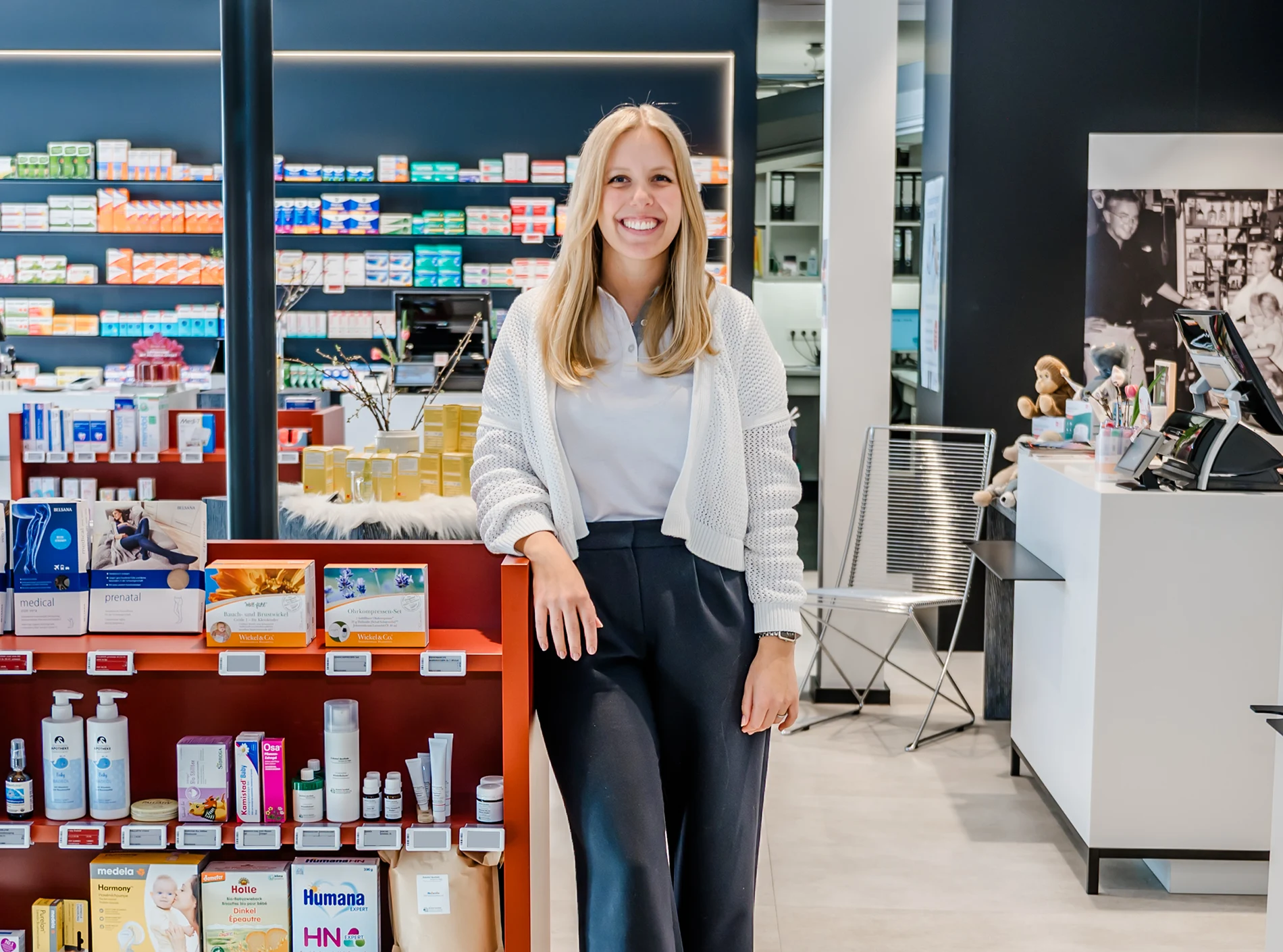 Eine lächelnde Frau steht in einer modernen Apotheke und lehnt an einem Regal mit Gesundheitsprodukten und Medikamenten. Bunte Verpackungen säumen die Regale, im Hintergrund ist der Apothekentresen zu sehen.